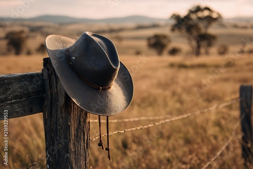 A weathered cowboy hat rests on a rustic wooden fence post in a sun-drenched rural landscape.