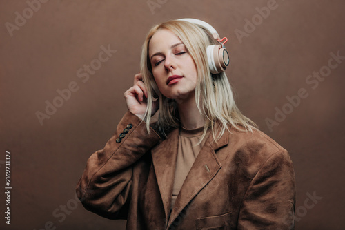 A stylish blonde woman wearing a brown jacket paired with modern headphones, standing against a rich brown background in shade Mocha Mousse