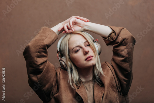 A stylish blonde woman wearing a brown jacket paired with modern headphones, standing against a rich brown background in shade Mocha Mousse