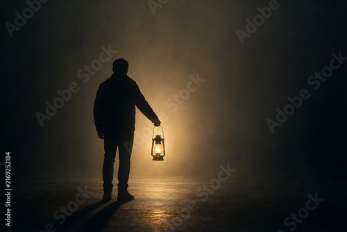 Silhouette man holding lantern in dark foggy night, glowing light illuminating mysterious misty surroundings