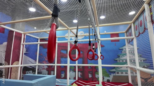 Indoor playground obstacle course featuring hanging red gymnastics rings and safety nets.