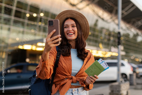 Happy woman holding passport with boarding passes while taking selfie or video calling with her smartphone outside airport terminal