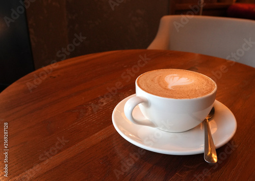 White cup of cappuccino with latte art on a wooden table. Warm cozy cafe photography. Ideal for coffee shop promotions, lifestyle blogs, or breakfast menu designs.