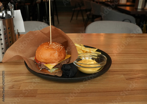 Craft cheeseburger in paper, fries, cheese sauce and black gloves on a plate. Wooden table in a blurred cafe interior. Ideal for fast food menus, pub advertising or food blogs.