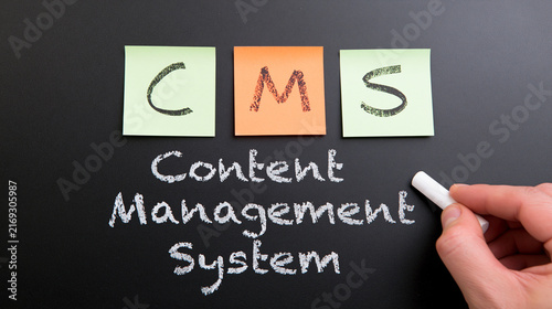 A hand writes Content Management System with chalk on a blackboard beneath sticky notes. This visual concept illustrates digital website administration and business software.