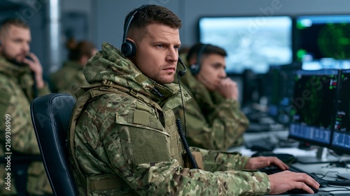 A focused soldier in camouflage uniform, wearing headphones, intently working on a computer in a dimly lit operations room.