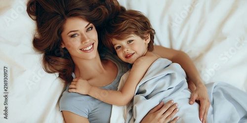 A tender moment captured between a mother and her child, both lying on a soft white bed. The mother has long, dark hair and a warm smile, while the child, wrapped in a blue blanket, beams with joy