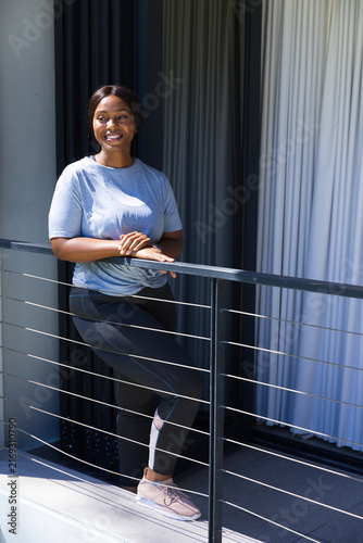 Фотография African American woman leaning on metal railing on apartment balcony, wearing te