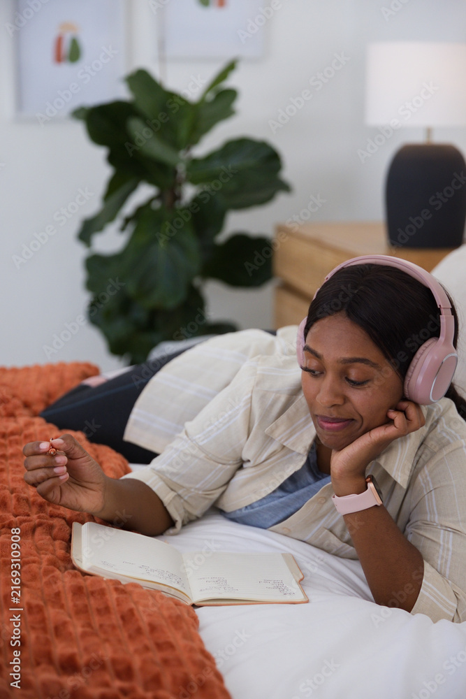 Fototapeta premium African adult woman lying on bed wearing pink headphones and writing in notebook in bedroom