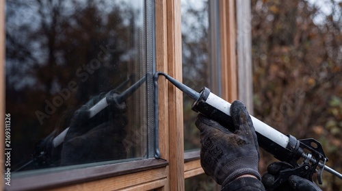 Close-up of a worker's hand in a protective glove applying grey silicone sealant to a wooden window frame using a caulking gun for weatherproofing and insulation.