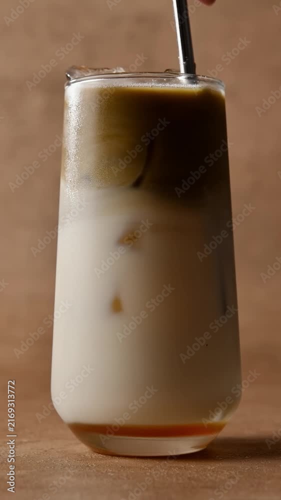 Hand stirs layered hojicha latte with straw, mixing roasted tea, oat ...