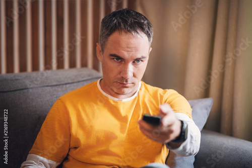 Middle-aged man in mustard-yellow t shirt sits on a grey sofa holding a TV remote; focused and serious expression. Wooden slats and beige curtains in the background