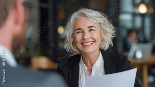Smiling mature business woman hr holding cv document at job interview. Happy mid aged professional banking manager or lawyer consulting client sitting at workplace in corporate office meeting.
