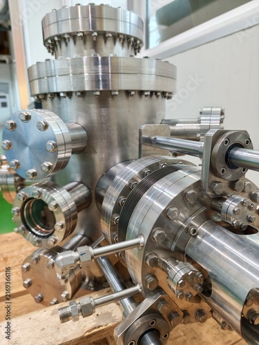 Fotografía A close-up shot of a high-vacuum chamber made of stainless steel with multiple conflat flanges, viewports, and linear motion feedthroughs resting on a wooden pallet inside a clean laboratory