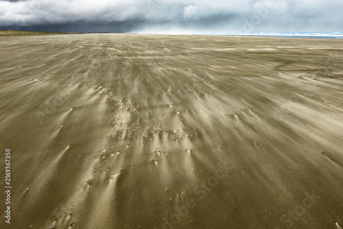 Distant storm clouds and windblown patterns with wind driven sand blowing across surface.