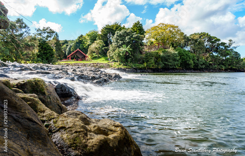 Waterfall over natural rock wier in Waipekakoura River outside Stone Store with red meeting hose structure on other side.