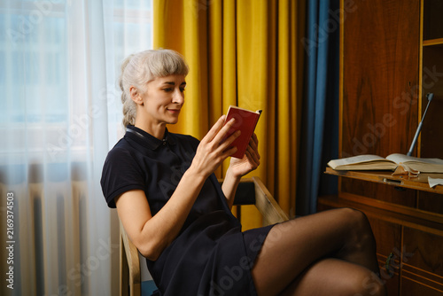 Woman with grey hair reading paper book in cozy room with bright curtains and wooden furniture