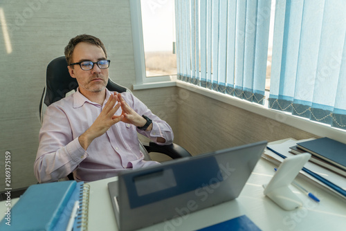 Businessman sitting at his office desk with his hands clasped in front of him, looking thoughtful and serious. Business meeting, waiting, or considering important decisions.