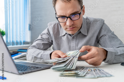 Fotografie Businessman carefully counting a large stack of dollar bills on his desk while working on a laptop