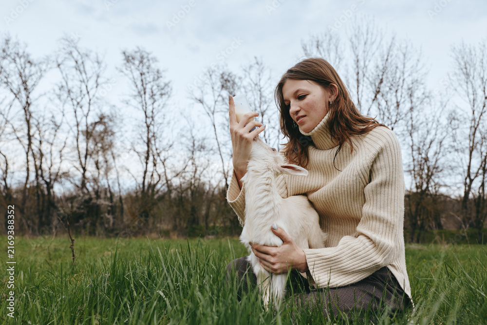 Naklejka premium Woman in a cozy sweater sits in a green meadow, embracing a fluffy goat kid, sharing gentle affection outdoors, conveying wholesome lifestyle moments and natural bond with animals.