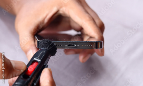 Asian man's hand cleaning a smartphone speaker grille with a small cleaning brush, also brushing around the charging port to remove dust, lint, and debris for better device maintenance and care.