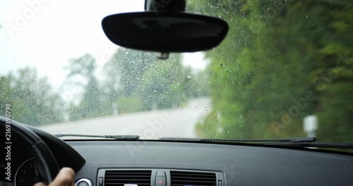 POV interior shot of a car with wet windshield driving a rainy country road. Gloomy weather, unrecognizable driver, rain drops