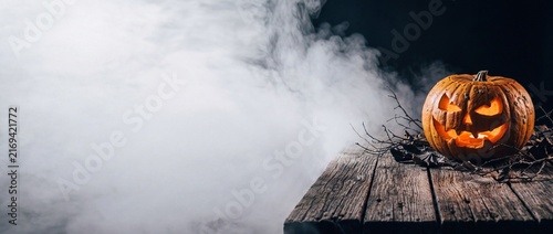 Menacing carved jack lantern resting on old rustic wooden table with bare branches surrounded by dense white smoke for halloween background