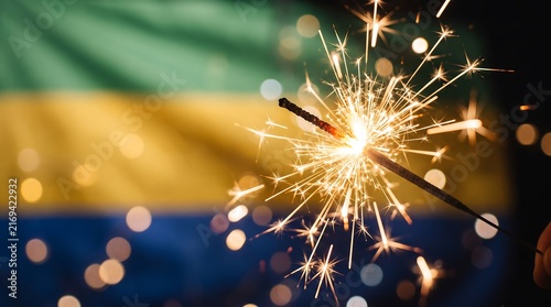 Burning sparkler macro shot against blurred Gabon flag background with warm bokeh light circles