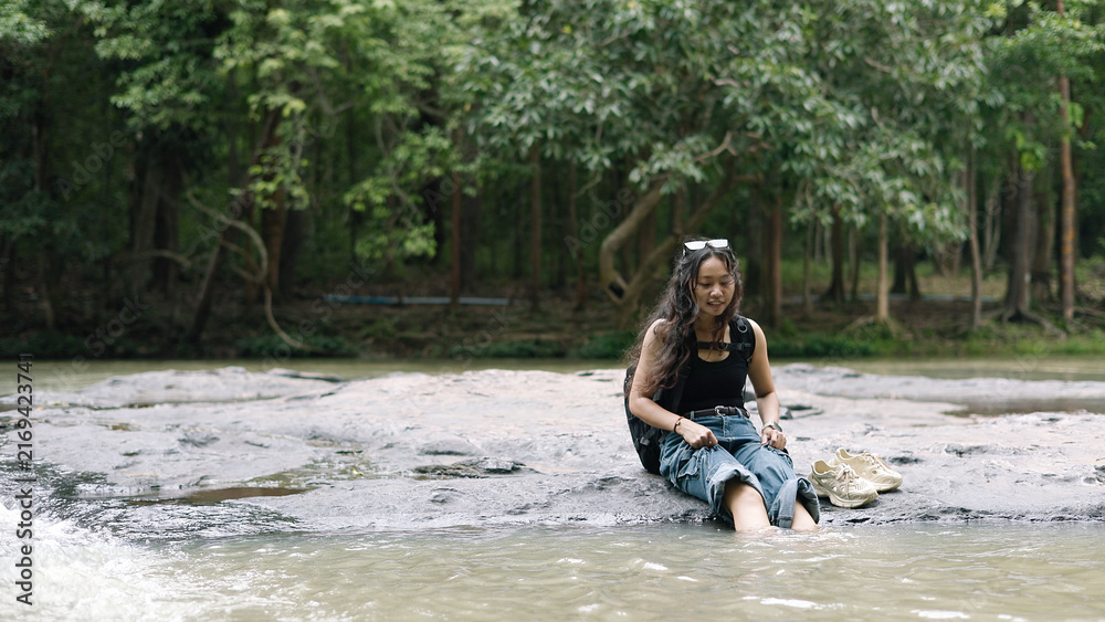 Fototapeta premium Young Female Backpacker Dipping Legs in River Stream to Relax