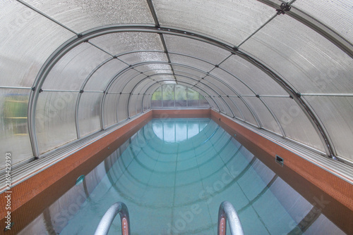 A long indoor swimming pool is inside a clear roof structure. The water is still. The pool is empty except for a few steps leading in. The sky is cloudy outside