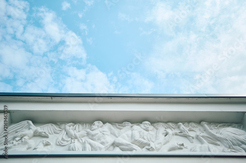 White carved frieze relief showing figures and winged creatures beneath building cornice, captured from low viewpoint with broad pale blue sky and soft clouds above, creating clean historical