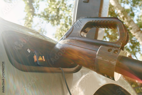 Electric vehicle charging plug connected to car inlet in outdoor setting, seen from low angle with bright sun flare, soft bokeh, reflective surfaces and detailed grip on connector handle