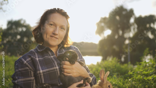 A farmer sits in the sunlight, holding rabbits in her hands. the woman with the rabbits, surrounded by greenery, under the warm sun.