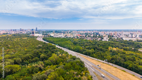 City skyline and forest view with road traffic seen from above. Aerial view showing a city with buildings and forest nearby. Roads have vehicles moving.