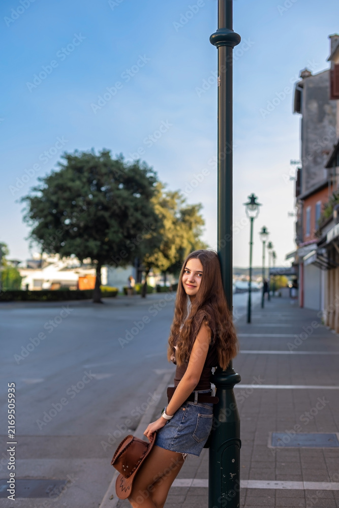 Fototapeta premium Young Woman Posing by Streetlamp with Cowboy Hat. Young woman with long brown hair posing beside a streetlamp in denim shorts and a brown top while holding a cowboy hat.