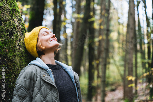 A young woman in a yellow beanie leans comfortably against a massive tree trunk in a quiet autumn forest. Relaxing nature connection concept emphasizes the grounding 
