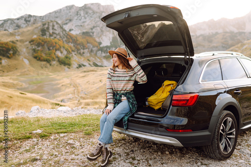 Bild auf Leinwand Woman in hat sits on edge of car's trunk, admiring mountain scenery
