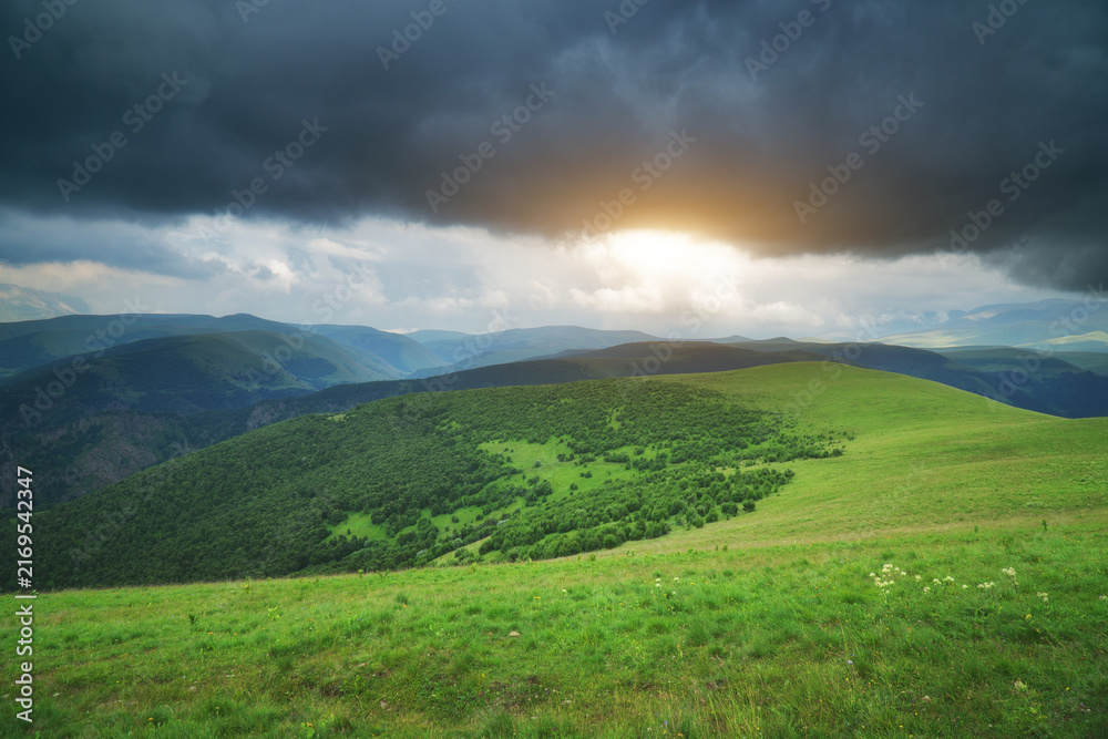 Fototapeta premium Bright sunset breaks through dark storm clouds over lush green hills and alpine meadows. Majestic mountain landscape