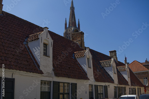 Row of traditional white historic houses with red tile roofs and dormers below a towering gothic church spire in Bruges, Belgium. Travel landmark.