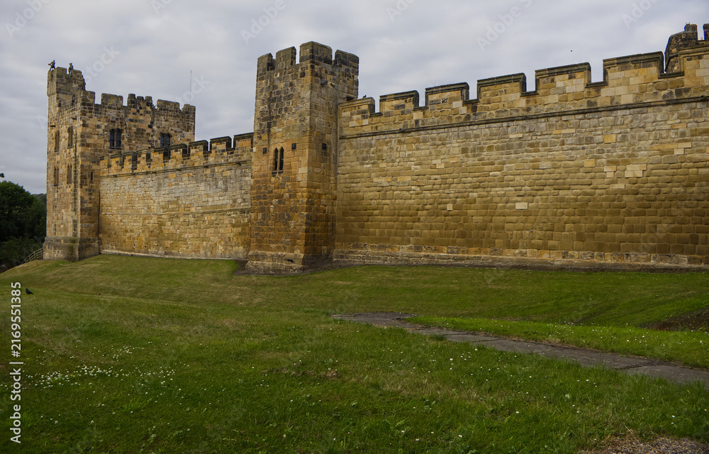 Obraz premium Majestic medieval stone castle fortress featuring ancient defense walls, battlements, and tall towers surrounded by green grass under a cloudy sky. Alnwick castle England, towers a very old castle