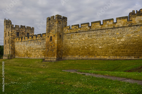Foto Majestic medieval stone castle fortress featuring ancient defense walls, battlements, and tall towers surrounded by green grass under a cloudy sky