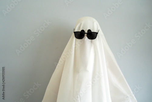 Cool ghost in a white costume wearing stylish black sunglasses, funny Halloween concept with minimalist background, spooky holiday, disguise and humor.