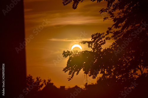 The golden sunset during the solar eclipse hides behind the silhouettes of trees and rooftops, creating a warm, golden sky and a peaceful evening atmosphere.