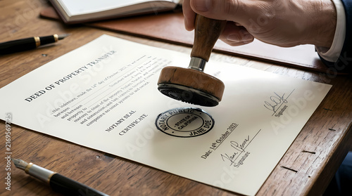 A business professional uses a wooden rubber stamp to certify an official legal deed of property transfer document on a wooden desk to finalize a real estate transaction between the two parties