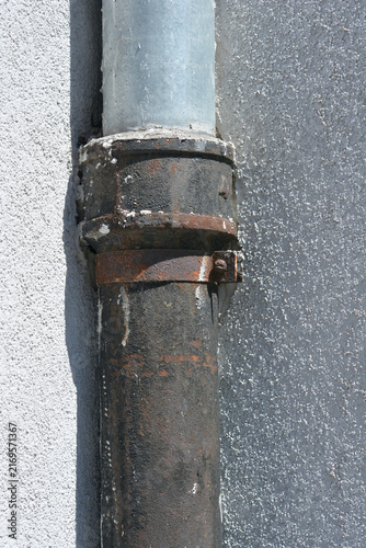Fotografie A close up view of an old rusted cast iron drainpipe connected to a galvanized steel pipe on a textured white facade wall