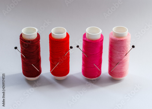 Four spools of sewing thread in burgundy, red, magenta and light pink with black headed sewing pins inserted into the thread, arranged on a clean light background.