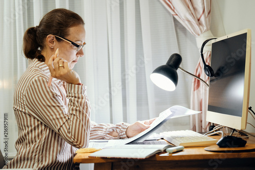 Mid-adult woman in glasses turning pages of documents at home desk with lamp and computer monitor while working on project in evening.