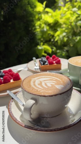 Close-up outdoor cappuccino with latte art and berry tarts on ceramic tableware, surrounded by green foliage and sunlight, cozy summer café atmosphere, lifestyle food photography.