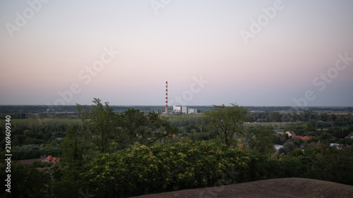 Canvastavla Tall red and white striped industrial chimney rises above power plant, viewed from distance over lush green canopy