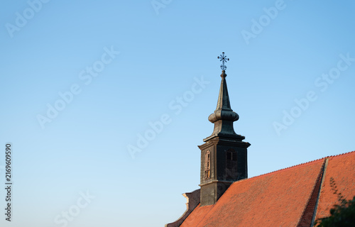 Historic Petrovaradin church spire with decorative metal cross rises above red tiled roof against clear blue sky. Novi Sad, Serbia